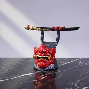 K&ouml;nnte beinhalten: Ein roter und goldener japanischer D&auml;monenmasken-St&auml;nder, der ein schwarzes Schwert h&auml;lt. Die Maske hat goldene Sto&szlig;z&auml;hne und ein detailliertes Design. Das Schwert hat einen goldenen Griff und rote Akzente. Der St&auml;nder steht auf einer schwarzen Marmoroberfl&auml;che.
