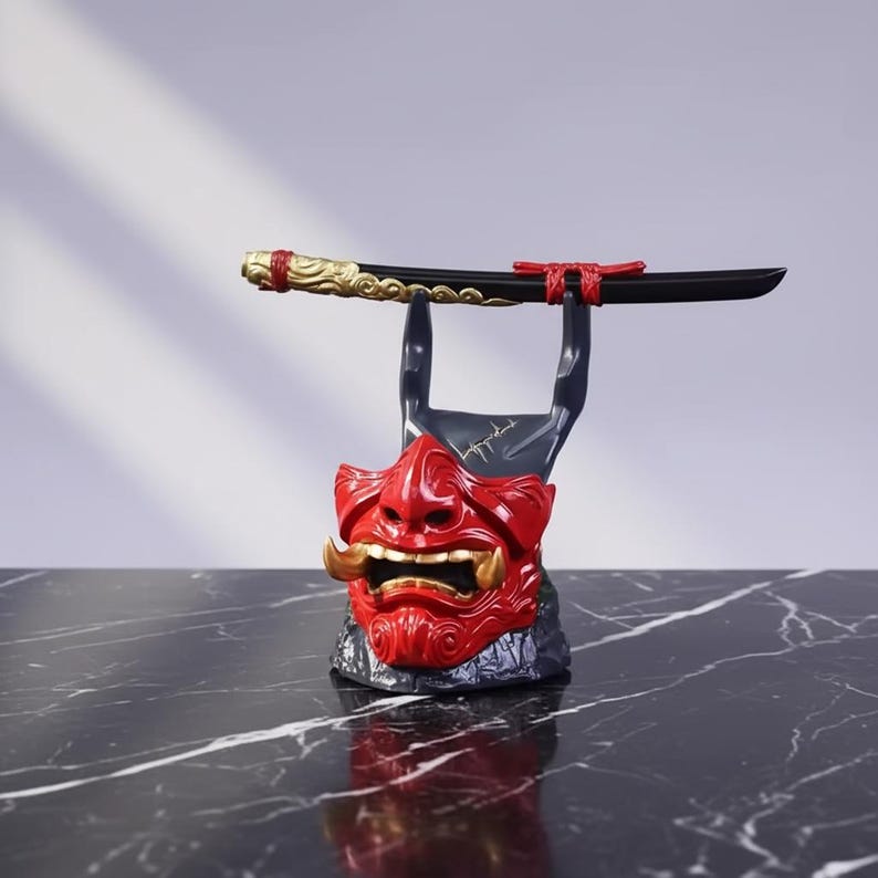 K&ouml;nnte beinhalten: Ein roter und goldener japanischer D&auml;monenmasken-St&auml;nder, der ein schwarzes Schwert h&auml;lt. Die Maske hat goldene Sto&szlig;z&auml;hne und ein detailliertes Design. Das Schwert hat einen goldenen Griff und rote Akzente. Der St&auml;nder steht auf einer schwarzen Marmoroberfl&auml;che.