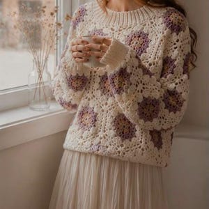 Puede incluir: Un suéter de ganchillo de manga larga con una base de color crema y motivos florales repetitivos en tonos lavanda, rosa suave y amarillo apagado. La prenda tiene un cuello redondo acanalado y puños acanalados, combinado con una falda larga plisada color crema.