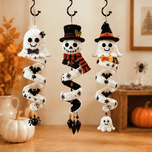 May include: Three hanging crochet ornaments featuring Halloween themes. The left is a white ghost with a purple bow, the center is a skeleton in a top hat and scarf, and the right is a ghost wearing a witch hat and orange bow, all with spiral tails.