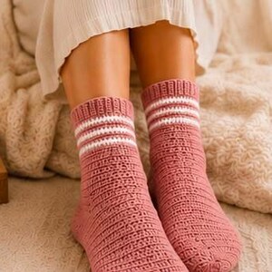 May include: A pair of dusty rose knitted socks featuring a ribbed cuff with two horizontal white stripes. The socks have a textured, vertical stitch pattern throughout the body and reach mid-calf, providing a consistent knit construction.