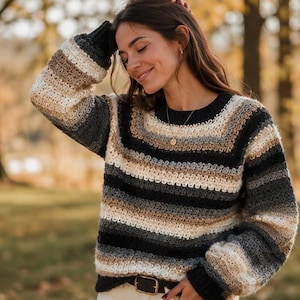 May include: A long-sleeved crochet sweater featuring horizontal stripes in shades of charcoal gray, cream, and tan. The garment has a round neckline with a ribbed collar, ribbed cuffs, and a ribbed hem, all constructed with a textured yarn pattern.