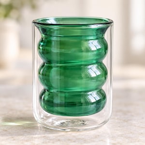 Może przedstawiać: Szklanka o podwójnych ściankach z żywą, szmaragdowozieloną warstwą wewnętrzną z trzema wyraźnymi, poziomymi, zaokrąglonymi wypukłościami. Zewnętrzna ścianka jest przezroczysta i gładka, tworząc cylindryczną sylwetkę z szeroką, otwartą krawędzią.