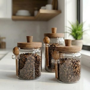 Könnte beinhalten: Drei durchsichtige Glasbehälter gefüllt mit gerösteten Kaffeebohnen, jeweils mit einem runden Holzdeckel und einem passenden Holzknauf. Jeder Behälter hat eine kleine Glasschlaufe an der Seite, die einen kleinen Holzlöffel zum Schöpfen hält.