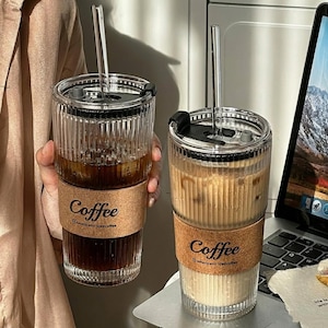 Könnte beinhalten: Zwei geriffelte Glastrinkbecher mit schwarzen Deckeln und klaren Glasstrohhalmen. Jeder Becher hat eine Korkmanschette, die mit dem Wort Coffee und dem Text Americano iced coffee bedruckt ist. Ein Glas enthält dunkle, das andere helle Flüssigkeit.