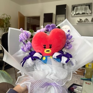 Könnte beinhalten: Ein roter, herzförmiger Plüschcharakter mit gelbem Gesicht und schwarzen Details, umgeben von lila Blumen und in weißes Papier gewickelt. Die Figur hat ein blaues und gelbes Outfit. Der Strauß ist mit einem lila Band gebunden.