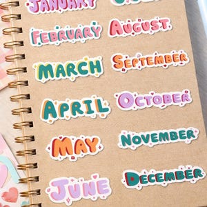 Puede incluir: Un cuaderno espiralado con pegatinas coloridas de los meses del año. Cada mes está escrito en una fuente divertida con un contorno blanco y elementos decorativos. Las pegatinas son de varios colores.