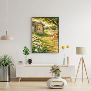 May include: A framed watercolor painting depicts a serene outdoor scene. A small white cottage with a window is covered in vines. A hammock hangs from a tree, and a picnic blanket rests on the grass. White and pink flowers are in the foreground.