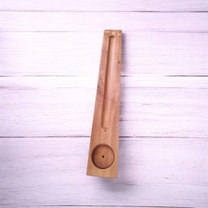 Puede incluir: Un porta incienso de madera de forma rectangular, con una hendidura circular en la base y una ranura central. El soporte de madera de color marrón claro está diseñado para sostener varillas de incienso. Mide unos 30 cm de largo.