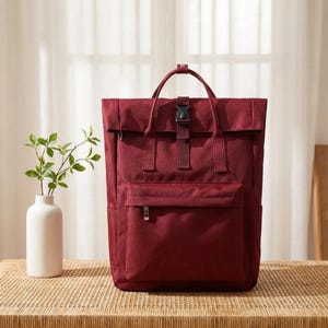 May include: A deep burgundy roll-top backpack featuring a rectangular silhouette, a front zippered pocket, two top carry handles, and a black plastic buckle closure centered on the front flap. The fabric has a matte, textured finish.