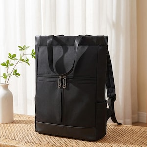 Può includere: Uno zaino rettangolare nero dotato di maniglia superiore, due spallacci e una tasca frontale con cerniera e due cursori in metallo argentato. La borsa presenta un design strutturato con base rinforzata e tasche laterali per riporre oggetti.