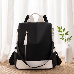 Könnte beinhalten: Ein schwarzer Rucksack mit weißen Verzierungen und Akzenten. Die Tasche verfügt über eine vertikale Reißverschlusstasche auf der Vorderseite mit goldenem Zipper, Seitentaschen, einen Tragegriff und verstellbare Schultergurte.