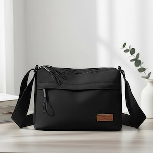 Op de afbeelding: Een zwarte crossbodytas met een lange, verstelbare schouderriem. De tas heeft een hoofdvak met ritssluiting en een voorvak. Een klein, rechthoekig bruin leren label met de merknaam staat op de voorkant. De tas staat op een licht oppervlak.