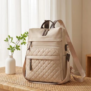 May include: A beige backpack featuring a quilted front pocket with a diamond pattern, a smooth upper section, dark brown zipper pulls, and adjustable shoulder straps. The bag includes a top handle and side pockets, constructed with a structured design.