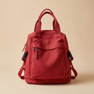 May include: A red canvas backpack featuring two top carry handles, a front zippered pocket with a matching red pull tab, and adjustable side straps secured with black plastic buckles. The bag has a structured base and a main zippered compartment.