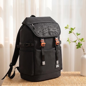 Könnte beinhalten: Ein schwarzer Rucksack mit einer oberen Klappe, die ein graues topografisches Linienmuster aufweist. Der Rucksack verfügt über zwei leuchtend orangefarbene Kunststoffschnallen auf der Vorderseite, verstellbare schwarze Seitengurte und einen kleinen rechteckigen Aufnäher mit weißem Text.