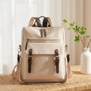 May include: A beige backpack featuring a large front pocket with two dark brown faux leather straps and gold-toned buckles. The bag includes a top handle, adjustable shoulder straps, and gold-toned zipper pulls on the main and front compartments.