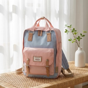 Peut inclure: Un sac à dos rose pastel et bleu clair doté d'un compartiment principal rectangulaire, d'une poche avant à rabat, de sangles en similicuir beige avec boucles métalliques, de deux poignées supérieures et d'une étiquette rectangulaire avec texte.