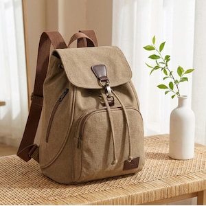 May include: A tan canvas backpack featuring brown fabric shoulder straps, a top flap with a metal hook closure, a drawstring main compartment, and a front zippered pocket. The design includes brown faux leather accents and a small rectangular patch.