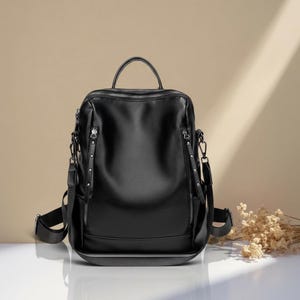 May include: Black leather backpack with a top handle and adjustable shoulder straps. The bag features multiple zippered compartments and silver-toned hardware. The design includes a structured shape and a sleek, modern aesthetic.