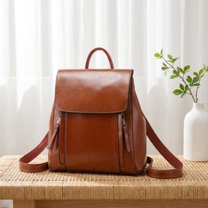 Peut inclure: Un sac à dos en cuir marron cognac lisse doté d'une fermeture à rabat, d'une poignée supérieure et de deux poches verticales zippées sur le devant. Le sac comprend des bretelles réglables avec des détails en cuir assortis et des ferrures.