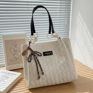 Può includere: Una borsa tote beige intrecciata con manici neri e dettagli ad anello color oro. La borsa presenta un fiocco marrone e una piccola etichetta con la scritta "CAMMER & MERA". La borsa è appoggiata su una superficie di legno.