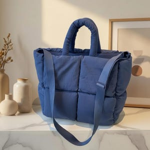 May include: A blue tote bag with a padded, quilted design. The bag has a top handle and a long, adjustable shoulder strap. The bag is made of a soft, durable material and is suitable for everyday use.