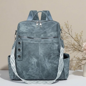May include: A denim-look backpack in a muted teal color. The bag features multiple zippered compartments, side pockets, and a patterned shoulder strap. The top has a handle and two zippers. The strap has a white and gray zigzag pattern.