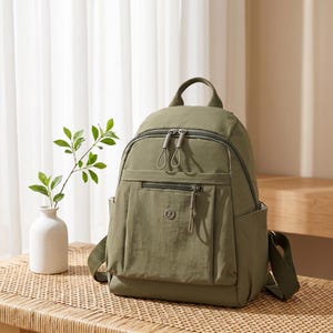 May include: An olive green backpack featuring a main zippered compartment, a smaller front zippered pocket with a circular logo containing the letter Z, two side pockets, a top handle, and adjustable shoulder straps with matching hardware.