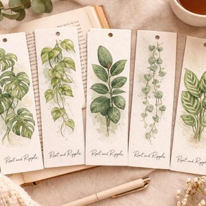 Könnte beinhalten: Fünf Aquarell-Lesezeichen mit botanischen Illustrationen verschiedener Pflanzen. Jedes Lesezeichen hat oben ein Loch und den Text "Root and Ripple" unten. Die Lesezeichen liegen auf einem Buch mit einem Stift und einer Tasse Tee.