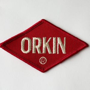 May include: Red embroidered patch in a diamond shape with the word "ORKIN" in cream-colored thread. The patch has a cream-colored border and a small circular logo at the bottom.