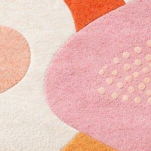 以下が含まれることがあります： ピンク、オレンジ、クリーム色の抽象的な形をした、柔らかく質感のあるラグのクローズアップ。ラグは、小さな隆起したドットと他の幾何学的な形を持つ大きなピンク色の形が特徴です。
