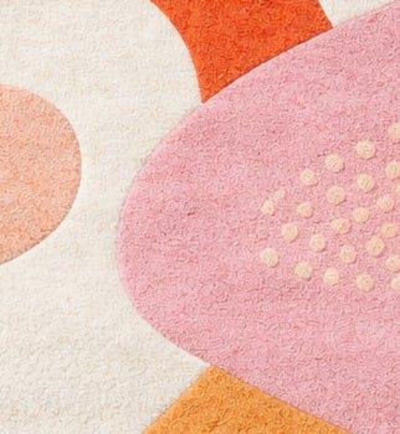 以下が含まれることがあります： ピンク、オレンジ、クリーム色の抽象的な形をした、柔らかく質感のあるラグのクローズアップ。ラグは、小さな隆起したドットと他の幾何学的な形を持つ大きなピンク色の形が特徴です。