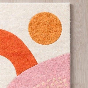 以下が含まれることがあります： 抽象的なデザインのクリーム色のラグの一部。ラグは、オレンジ色の円、オレンジ色のアーチ、小さな明るい色のドットが付いたピンク色の曲線形状が特徴です。 ラグは、質感のある豪華な外観をしています。