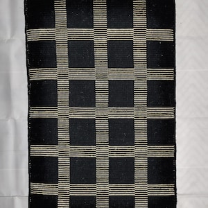 May include: A rectangular black and cream-colored woven rug with a grid pattern. The rug features large black squares separated by cream-colored horizontal and vertical stripes. The rug is likely made of natural fibers.