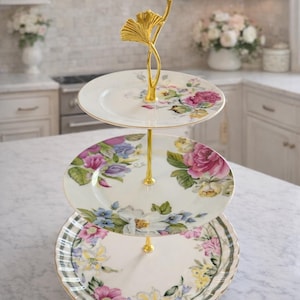 May include: A three-tiered serving stand with floral patterned plates. The plates are white with pink, purple, and blue flowers. The stand has a gold-colored metal handle with a leaf design. The stand is on a marble countertop.