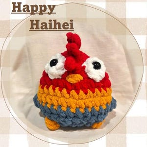 Puede incluir: Un pollo de ganchillo hecho a mano en rojo, amarillo y azul. El pollo tiene grandes ojos blancos con pupilas negras y una cresta roja. El texto "Happy Haihei" está en la parte superior de la imagen.