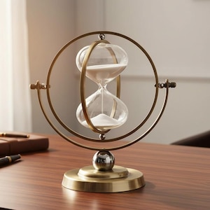 May include: A decorative hourglass with a clear glass bulb filled with white sand, set within a gold-coloured metal frame. The hourglass rests on a round base with a spherical accent. A stylish desk accessory.