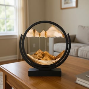 May include: A decorative sand art piece featuring a black circular frame with a rectangular base. Inside the frame, layers of sand create a mountain landscape. The sand is a light brown color, and the frame is made of metal.