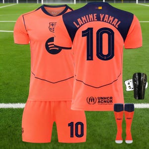 May include: An orange football kit, including a jersey with "LAMINE YAMAL" and the number "10" on the back. The kit also features shorts, socks, and black shin guards. The jersey has navy blue detailing around the collar and shoulders.