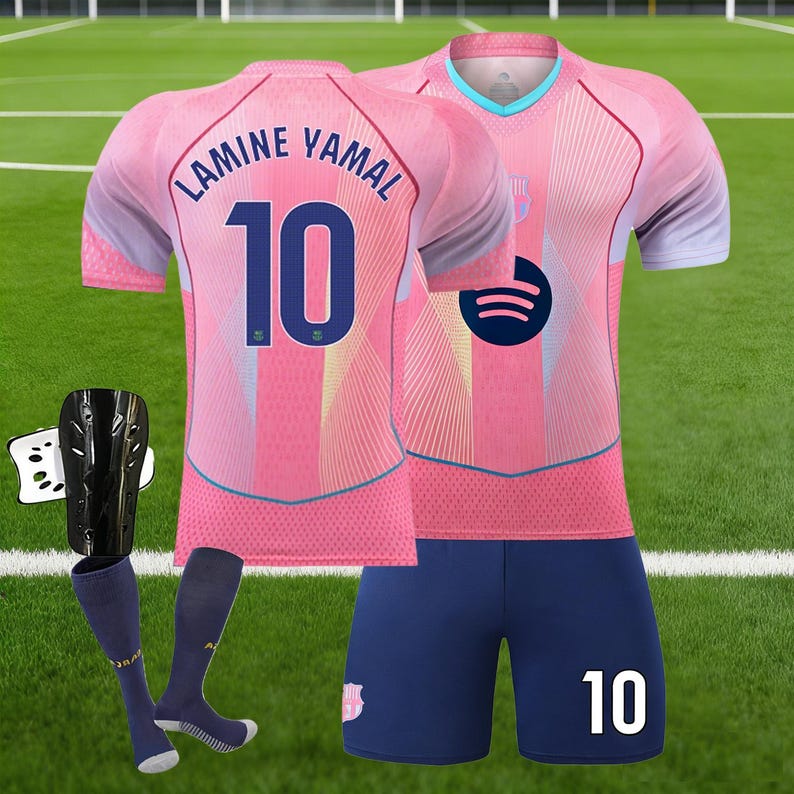May include: A pink soccer uniform set with the name "LAMINE YAMAL" and the number "10" on the back of the jersey. The set includes a jersey, shorts, and socks. The jersey has a Spotify logo. Also included are black shin guards.