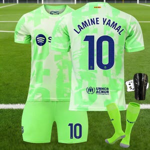 Puede incluir: Un uniforme de fútbol verde lima con el nombre "LAMINE YAMAL" y el número "10" en azul marino. El conjunto incluye pantalones cortos y calcetines a juego, además de espinilleras negras. La camiseta tiene el logo de Spotify.