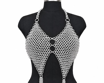 May include: A silver-colored chainmail halter top constructed from interlocking metal rings. The garment features a pointed, triangular design on each side, connected by central rings, with a halter neck strap and small dangling chain tassels.