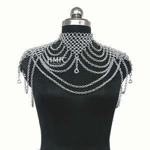 K&ouml;nnte beinhalten: Ein silberfarbenes Kettenhemd-Schulterst&uuml;ck mit hohem Kragen und mehreren drapierten Kettenlagen, die &uuml;ber die Schultern und die Brust verlaufen. Das St&uuml;ck zeigt den Text HMFC ARMOR in wei&szlig;em Druck auf dem vorderen Mittelteil.