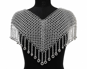 May include: A silver chainmail shoulder piece displayed on a black mannequin. The chainmail is made of small, interlocking metal rings, forming a draped, V-neck design with dangling fringe.