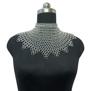May include: A silver chainmail collar, displayed on a black mannequin. The collar is made of small, interlocking metal rings, forming a protective neck covering. The design features a high, close-fitting neck and a scalloped edge.