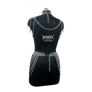 K&ouml;nnte beinhalten: Ein silberfarbenes Kettenhemd-K&ouml;rpergeschirr mit Halsband, Schulterdrapierungen und einem H&uuml;ftg&uuml;rtel mit mehreren herabh&auml;ngenden Kettenstr&auml;ngen. Das St&uuml;ck zeigt den Text HMFC ARMOR in Wei&szlig; auf der Mitte des schwarzen Schaufensterpuppen-Torsos.