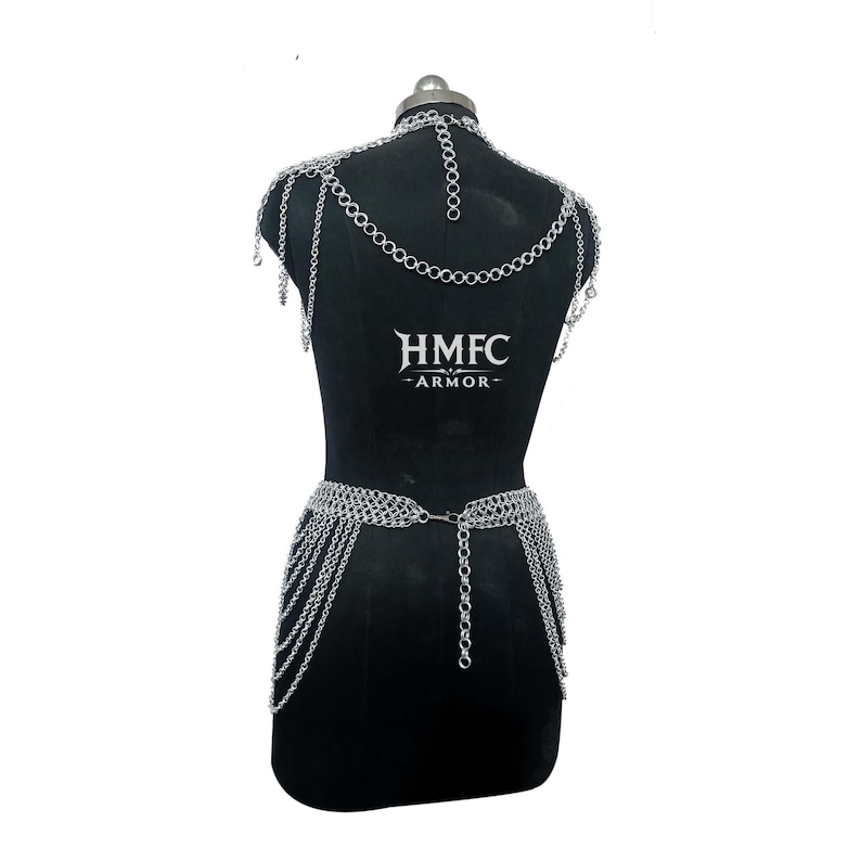 K&ouml;nnte beinhalten: Ein silberfarbenes Kettenhemd-K&ouml;rpergeschirr mit Halsband, Schulterdrapierungen und einem H&uuml;ftg&uuml;rtel mit mehreren herabh&auml;ngenden Kettenstr&auml;ngen. Das St&uuml;ck zeigt den Text HMFC ARMOR in Wei&szlig; auf der Mitte des schwarzen Schaufensterpuppen-Torsos.