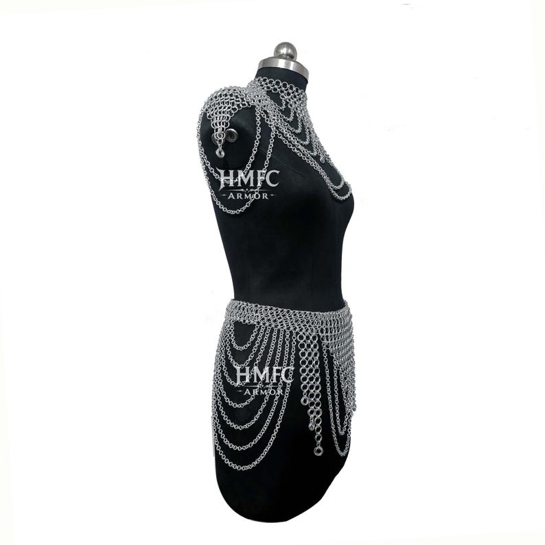 K&ouml;nnte beinhalten: Ein silberfarbenes Kettenhemd-K&ouml;rpergeschirr mit hohem Kragen, Schulterklappen und einem mehrlagigen, drapierten Taillenrock. Die Metallglieder bilden komplexe Muster &uuml;ber den Oberk&ouml;rper. Der Text HMFC ARMOR ist auf der Brust und H&uuml;fte sichtbar.