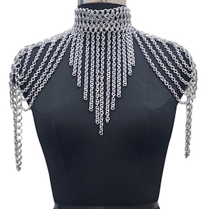 Puede incluir: Una pieza de hombro de cota de malla plateada con cuello alto. La cota de malla está hecha de pequeños anillos de metal entrelazados. El diseño incluye cadenas en cascada que caen sobre los hombros y el pecho.